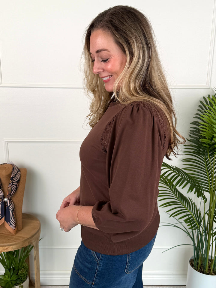 A woman wearing a brown long-sleeve top with lace-up puff sleeves and blue jeans, standing in a bright room with a white paneled wall, a potted plant, and a small wooden side table.
