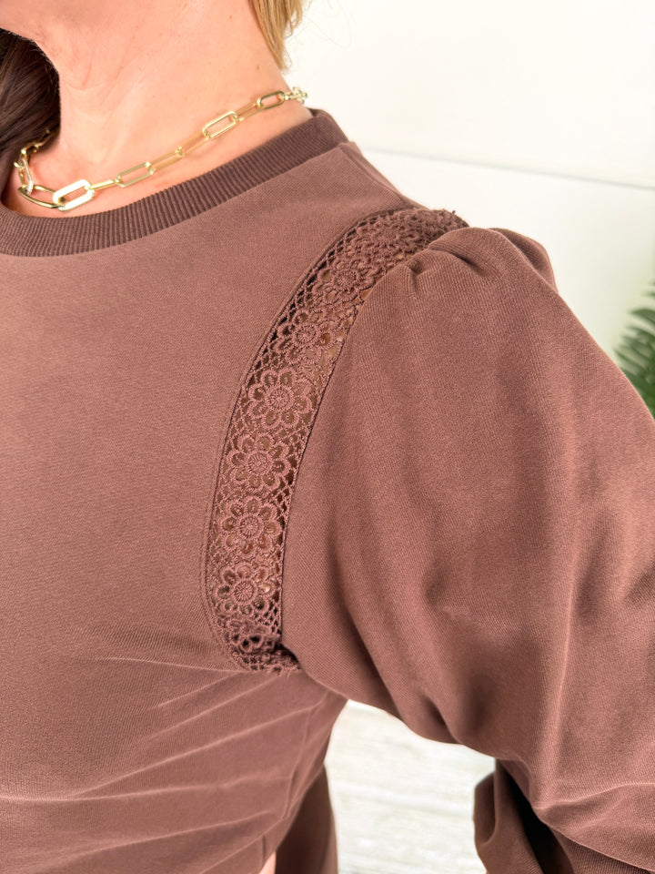 Close-up of a person wearing a brown long-sleeve top with a lace panel detail across the upper chest and shoulder, paired with a gold chain necklace.