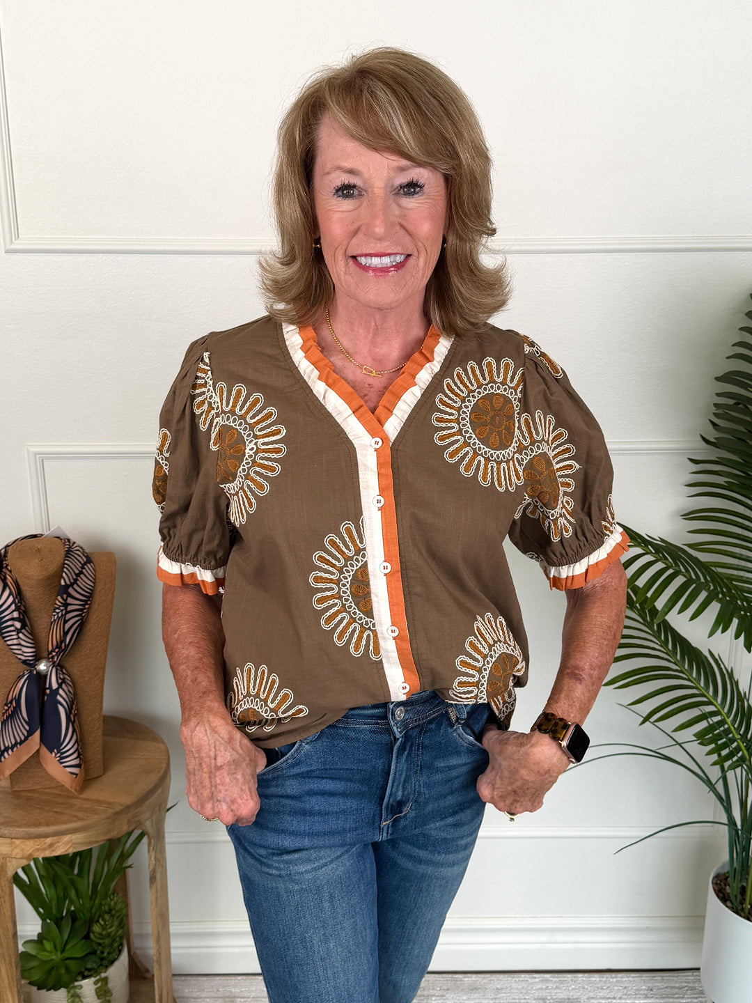 A smiling woman with blonde hair stands indoors wearing a brown short-sleeve blouse with orange and white sunflower-style patterns, paired with blue jeans. She is posed in front of a white paneled wall with potted plants and a wooden side table visible.