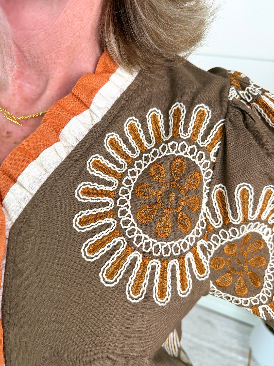 Close-up of a brown blouse with white and orange embroidered sunburst patterns, worn by a person with blonde hair and a gold necklace.