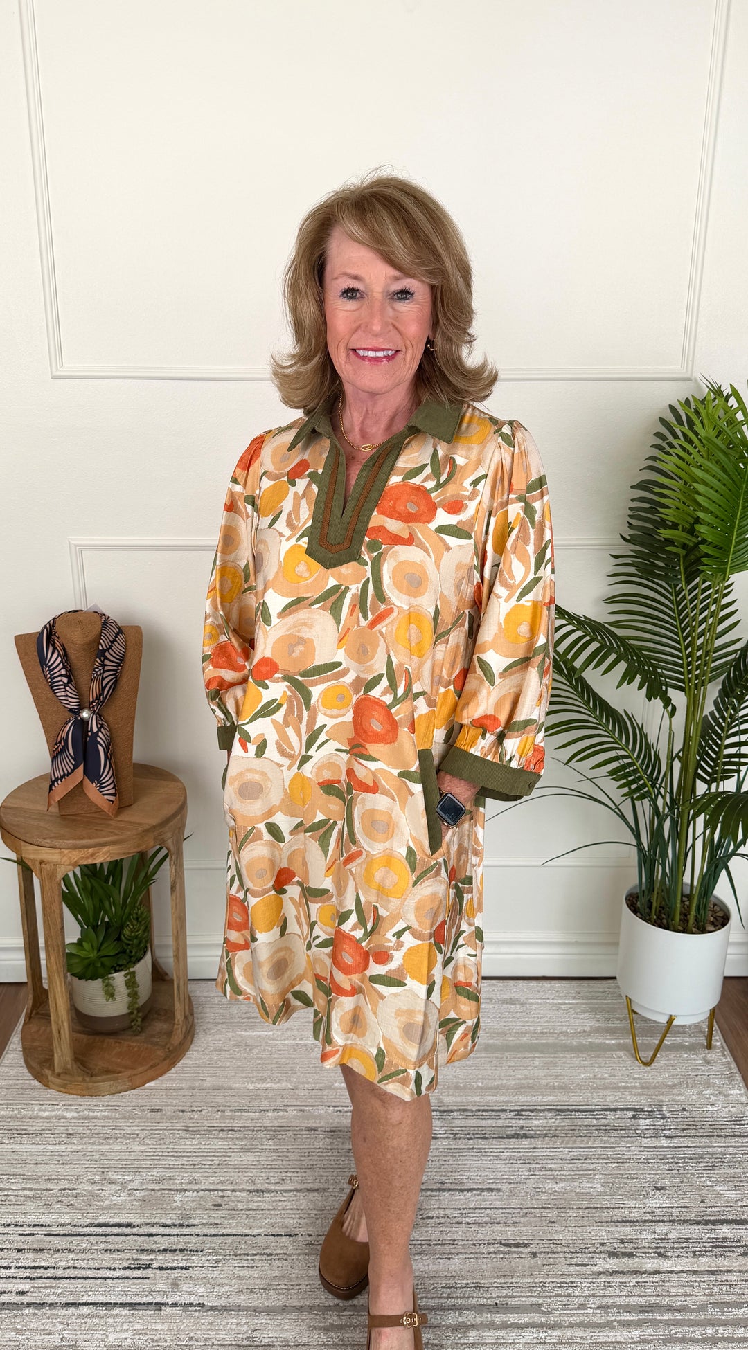 A woman modeling a short-sleeve collared dress with a retro orange, yellow, and green floral pattern, standing in a room with a white paneled wall, a potted plant, and a small wooden side table.