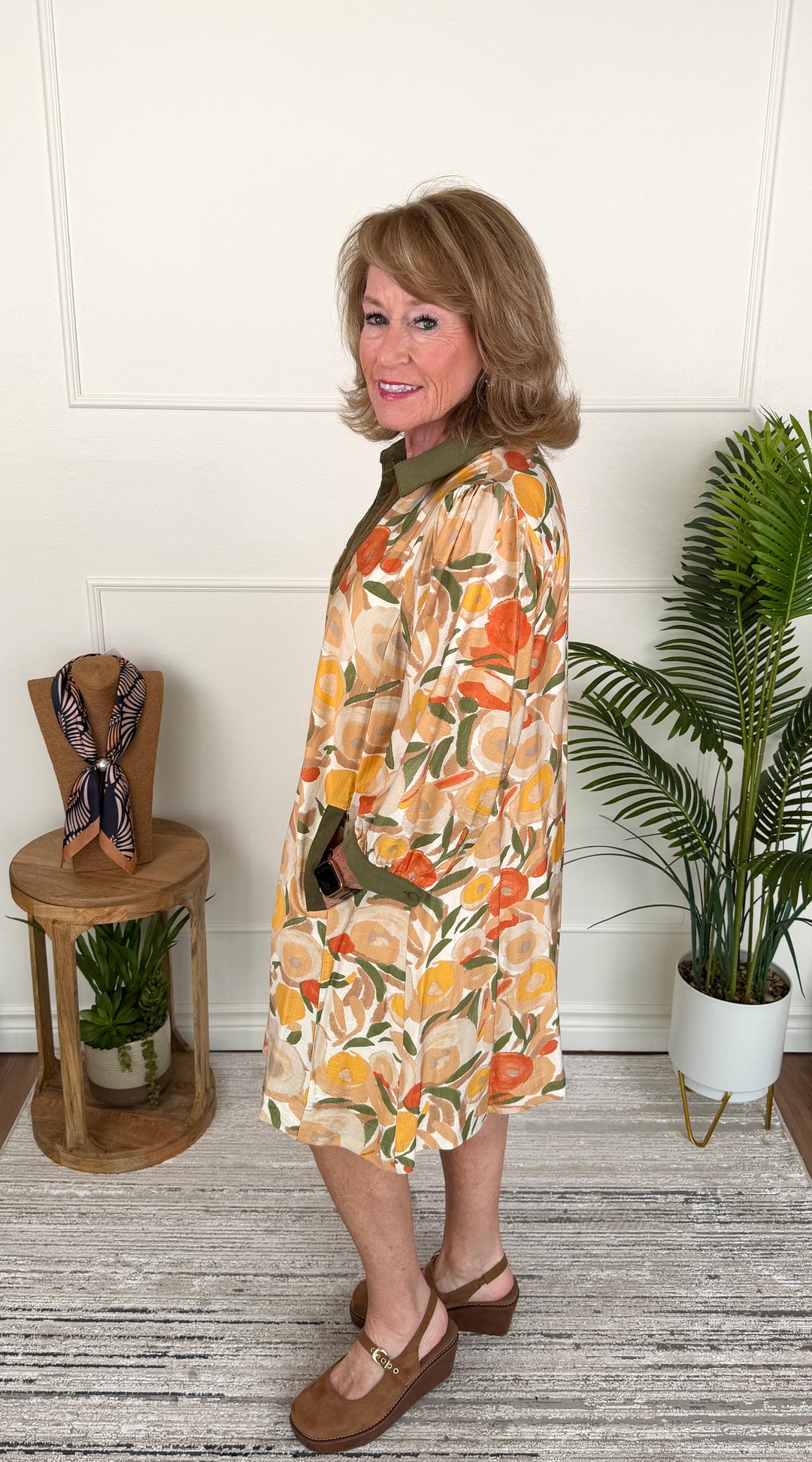 A woman modeling a knee-length floral dress with a green collar and green trim, standing in a styled indoor room with a white paneled wall, wooden floor, potted plant, and small side table.