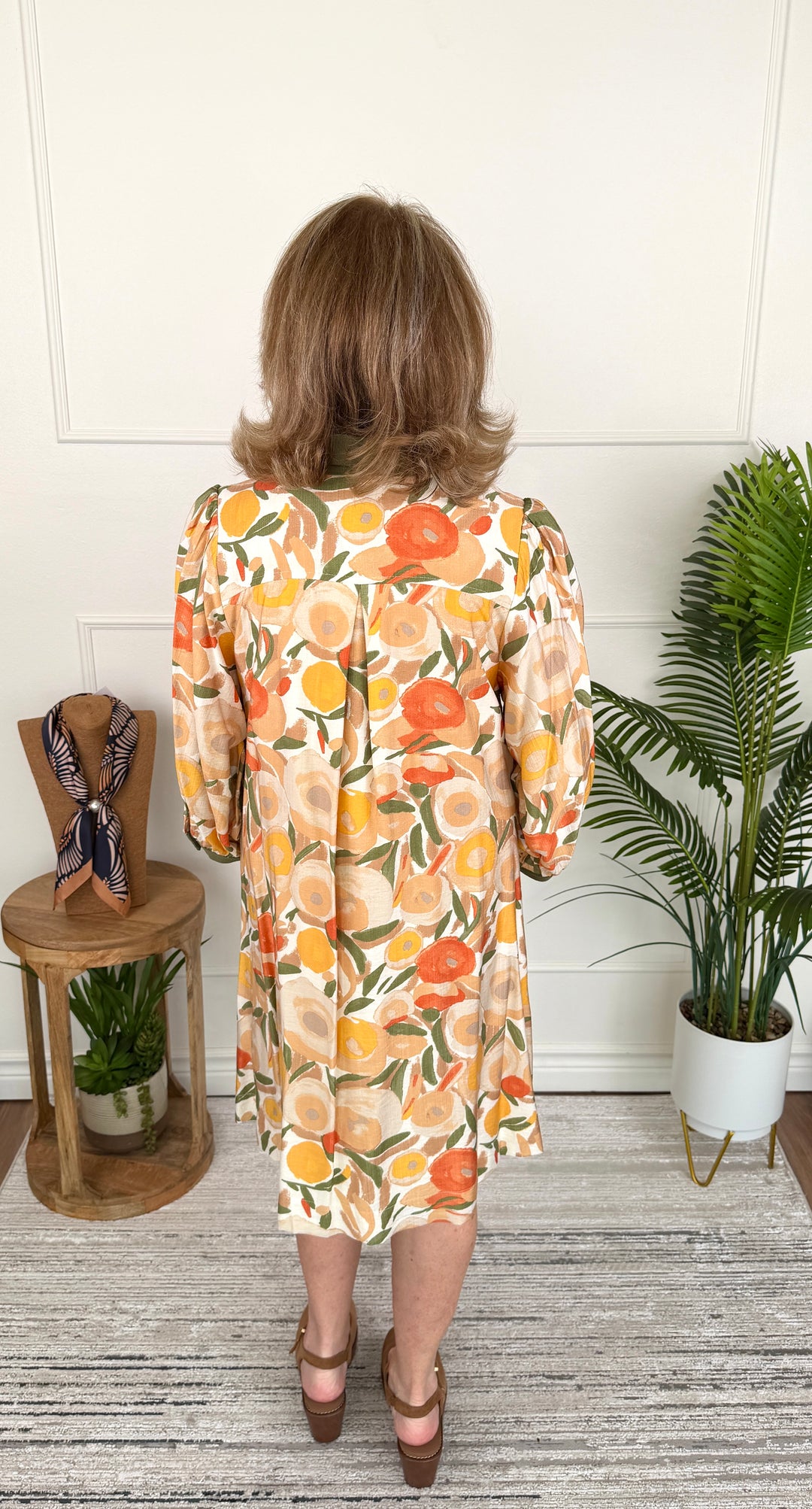 A woman viewed from behind wearing a long-sleeve midi dress with a vibrant orange, yellow, and green citrus fruit print on a cream background. She is standing in a room with a white paneled wall, a gray wood floor, a small wooden side table with a scarf, and a potted palm plant.