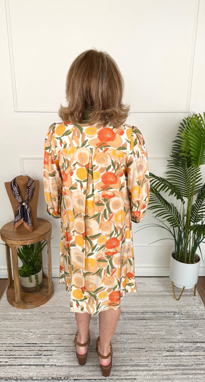 A woman viewed from behind wearing a long-sleeve midi dress with a vibrant orange, yellow, and green citrus fruit print on a cream background. She is standing in a room with a white paneled wall, a gray wood floor, a small wooden side table with a scarf, and a potted palm plant.