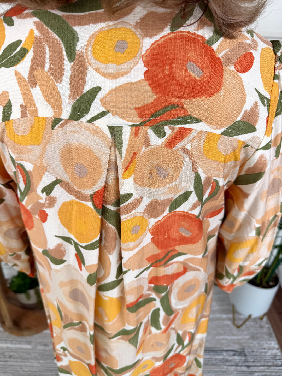 A close-up of a person wearing a short-sleeve top with a retro floral print in orange, yellow, beige, and green on a cream background.