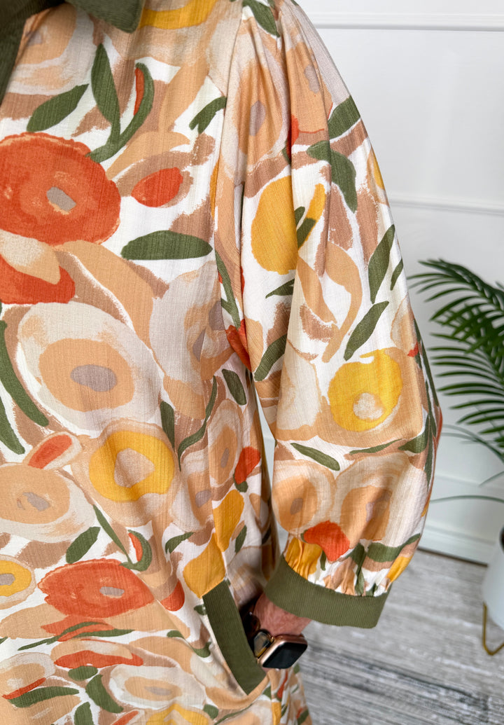 A close-up of a lightweight fabric garment featuring a retro floral pattern in orange, yellow, and green on a cream background, with an olive green cuff.