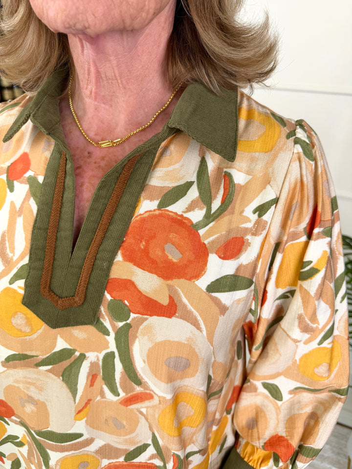 A close-up of a woman wearing a vintage-style floral blouse with an olive green collar and V-neck trim, featuring a warm orange, yellow, and beige floral pattern.