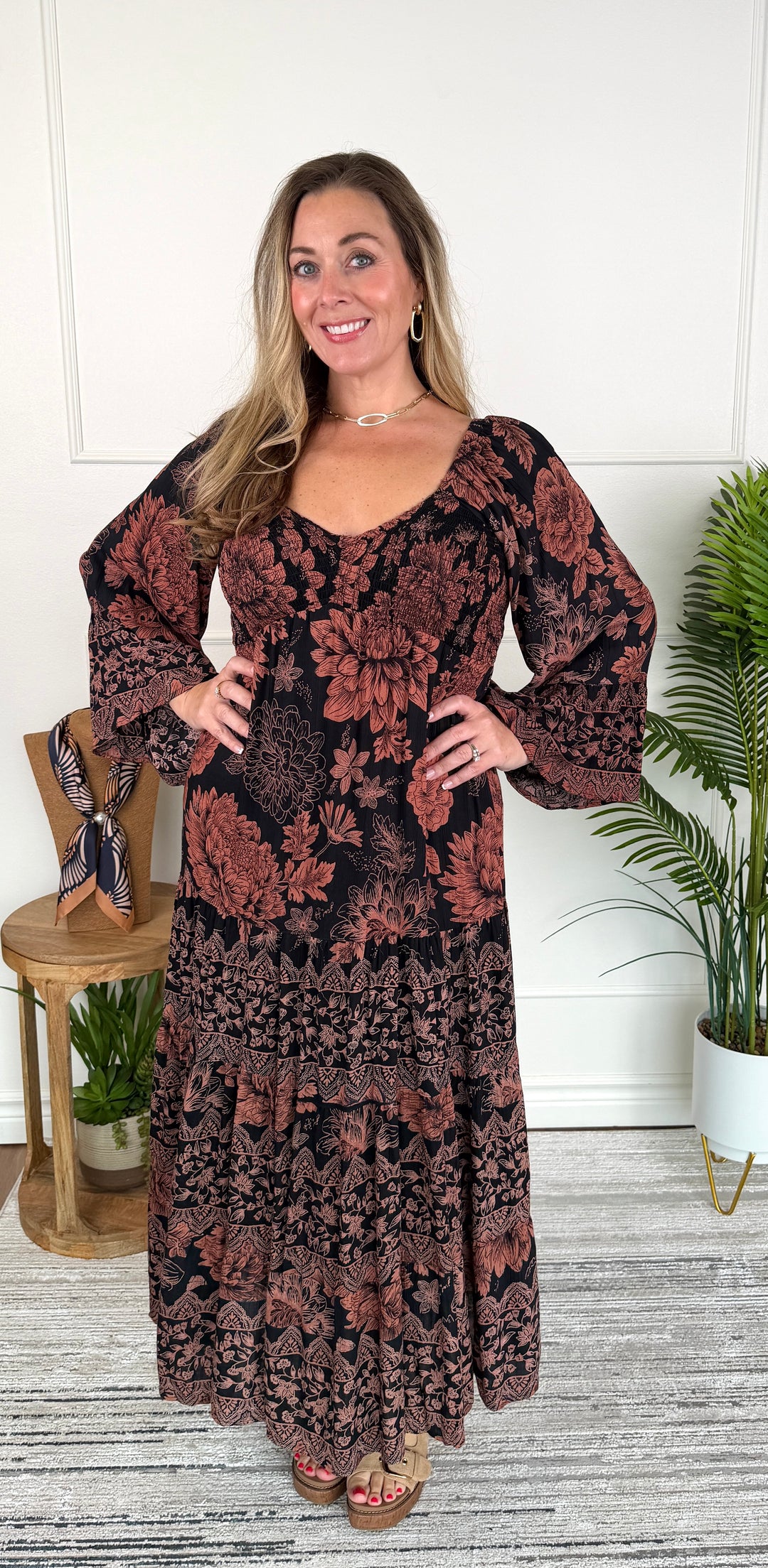 A woman modeling a long-sleeved maxi dress with a rust-brown and black floral pattern, standing in a brightly lit room with a white wall, gray wood floor, and potted plants.