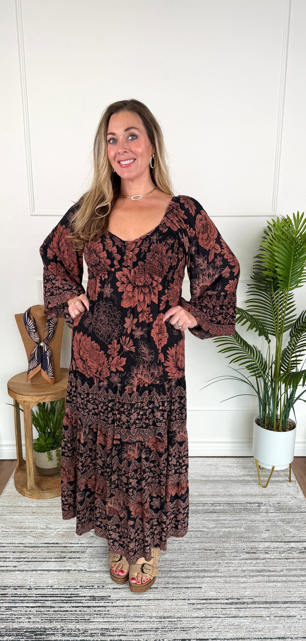 A woman modeling a long-sleeve bohemian maxi dress with a dark brown and rust floral pattern, standing in a room with a white paneled wall, a wooden chair, and potted plants.