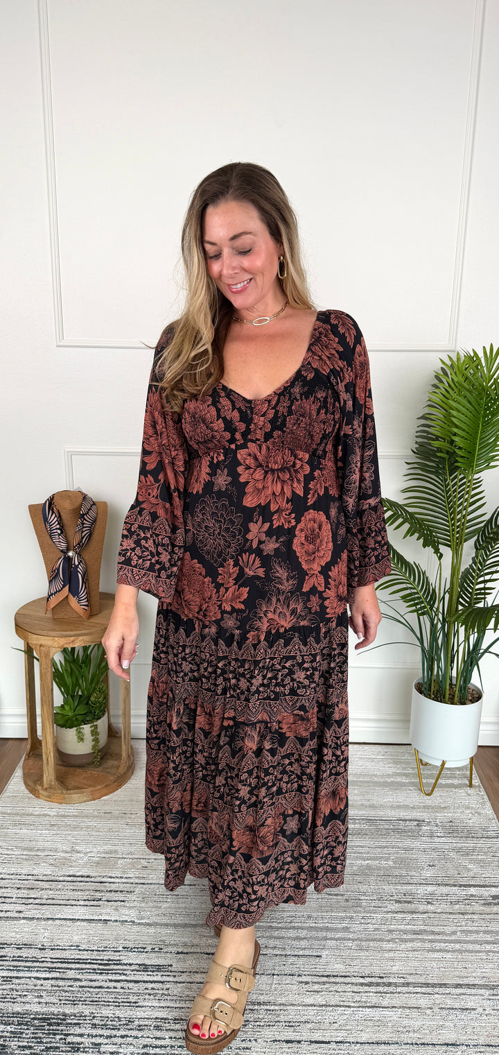A woman modeling a long-sleeve, tiered maxi dress with a dark floral pattern in rust red and black, standing in a styled indoor room with a potted plant and a small wooden side table.