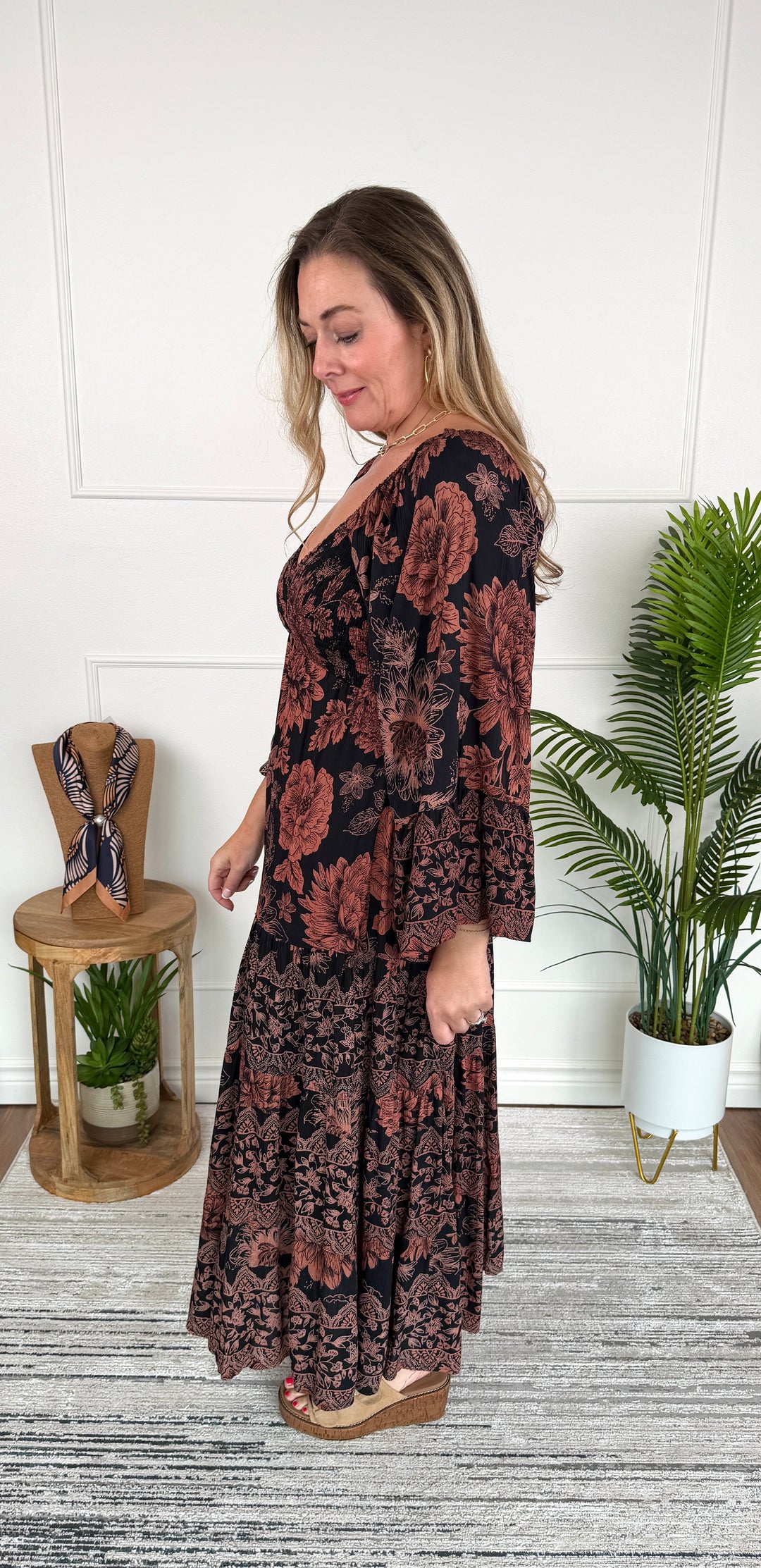 A woman modeling a long-sleeve maxi dress with a dark floral pattern, standing in a room with a white paneled wall, a potted plant, and a small wooden side table.
