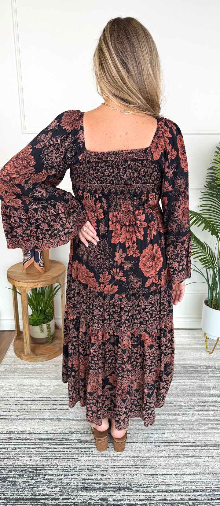 A woman viewed from the back wearing a long-sleeve bohemian-style maxi dress with a smocked bodice and tiered skirt in a dark brown and rust floral pattern, paired with brown wedge sandals.