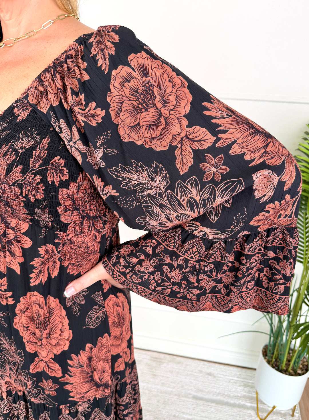 A close-up of a woman wearing a black dress with a large dusty rose floral print and ruffled bell sleeves, standing indoors next to a potted plant.