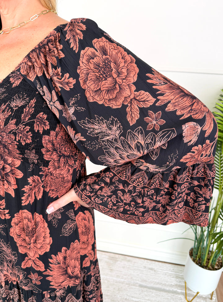 A close-up of a woman wearing a black dress with a large dusty rose floral print and ruffled bell sleeves, standing indoors next to a potted plant.