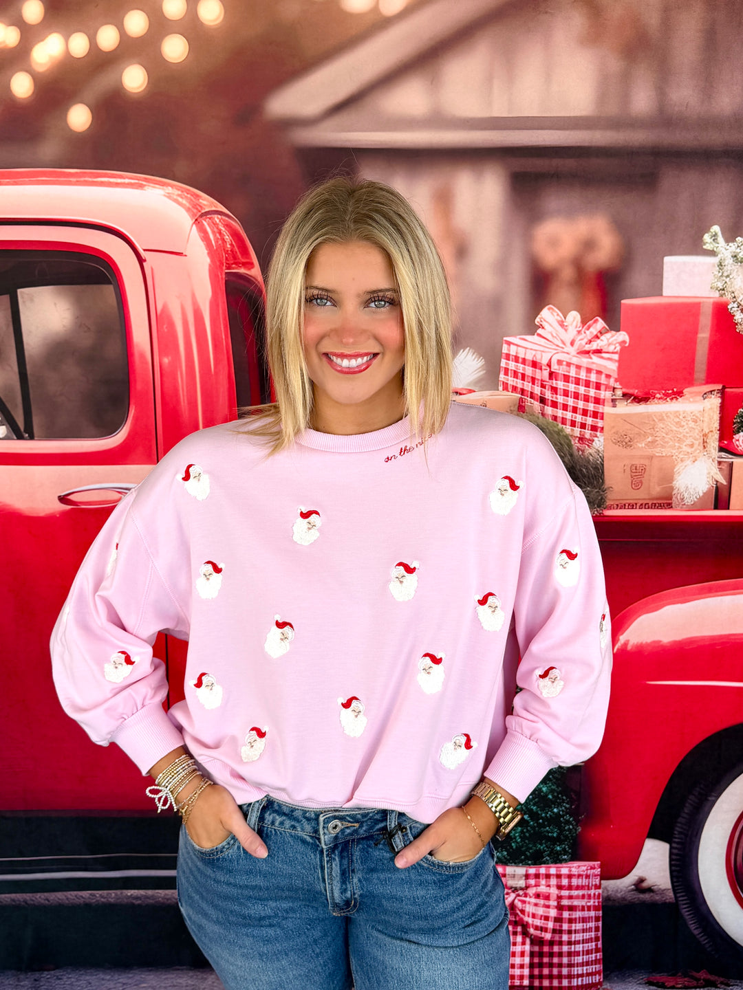 A smiling blonde woman wearing a light pink crewneck sweatshirt with a repeating Santa Claus print, paired with blue jeans, standing in front of a red vintage pickup truck decorated with holiday gifts.