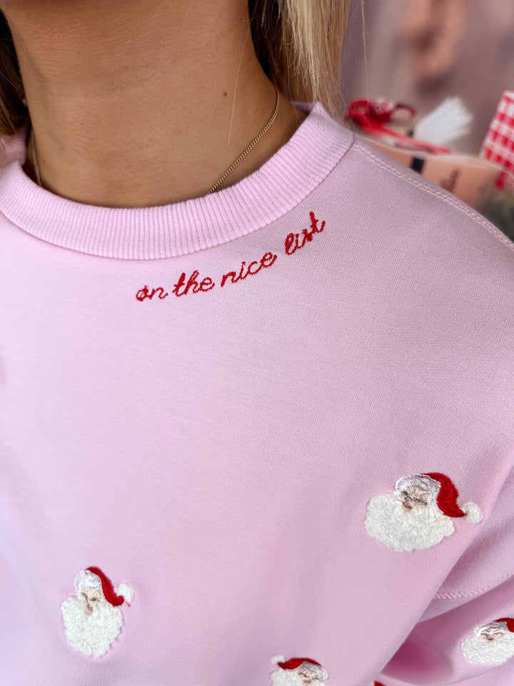 A close-up of a person wearing a light pink crewneck sweatshirt with red embroidered text reading 'on the nice list' and small white Santa Claus patches.