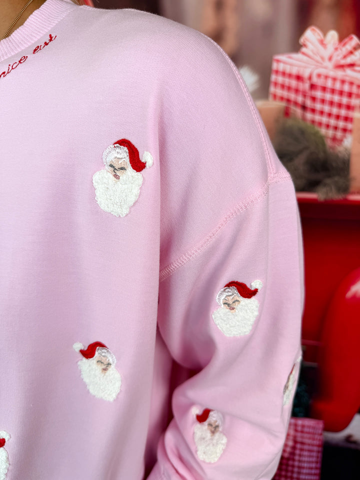 A close-up of a light pink crewneck sweatshirt featuring embroidered Santa Claus faces with white beards and red hats, worn by a model in a festive holiday setting.