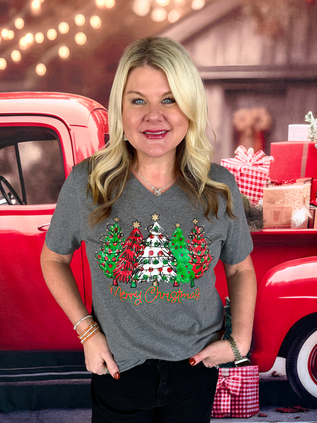 A woman wearing a gray graphic t-shirt with a Christmas tree design and 'Merry Christmas' text, standing in front of a red pickup truck decorated for the holidays.