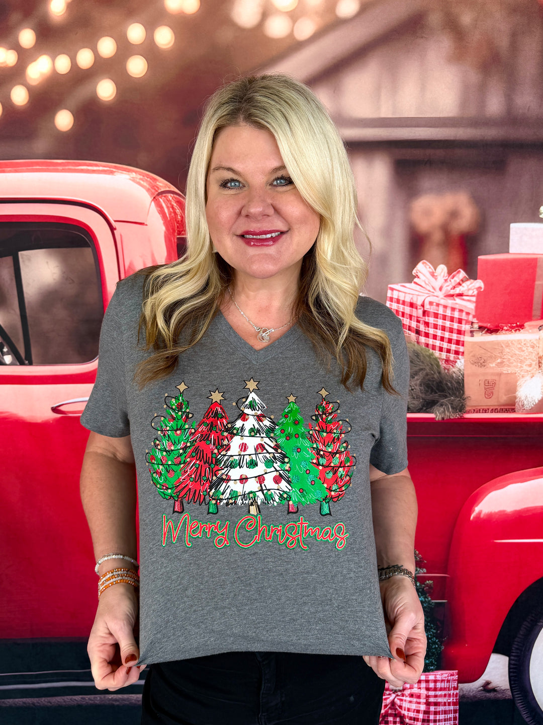 A woman wearing a heather gray V-neck t-shirt with a colorful Christmas tree graphic and 'Merry Christmas' text, posed in front of a red vintage truck with holiday decorations.