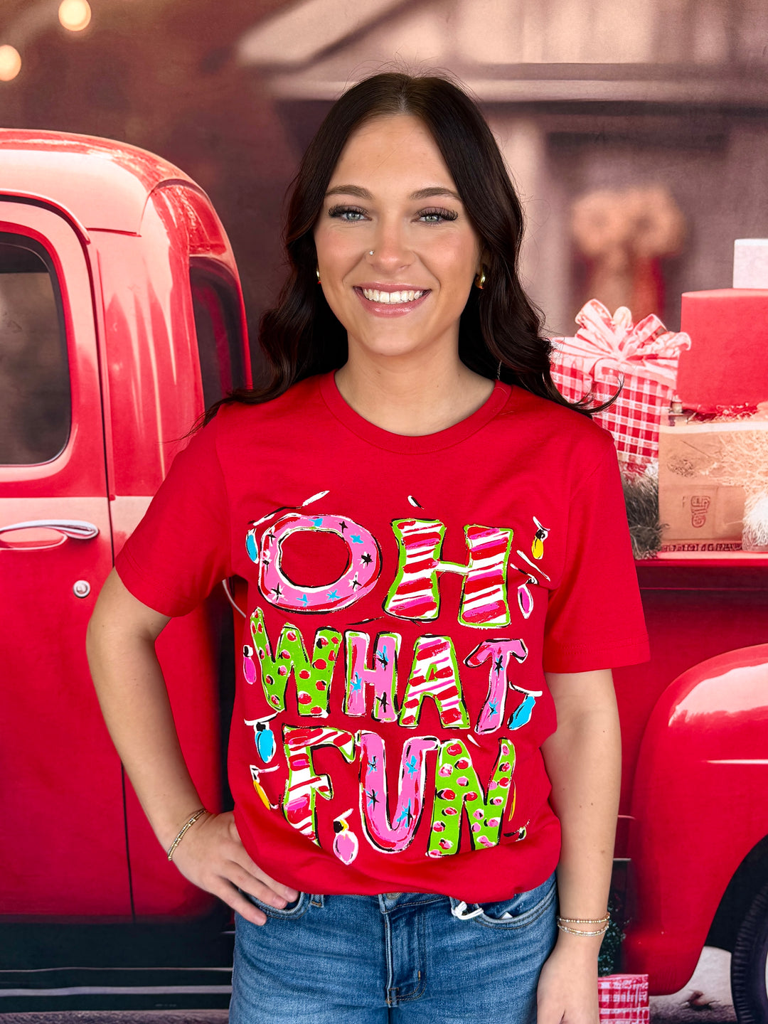 A woman smiling and wearing a bright red graphic t-shirt that reads 'OH WHAT FUN' in colorful, playful lettering. She is standing in front of a vintage red pickup truck with holiday decorations in the background.