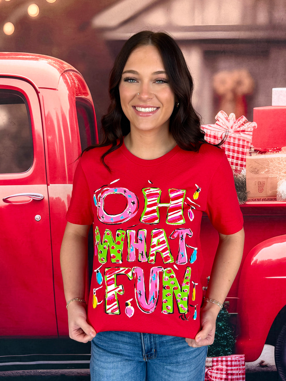 A woman wearing a bright red graphic t-shirt with the text 'OH WHAT FUN' in colorful, playful lettering, posed in front of a red vintage pickup truck with holiday decorations.
