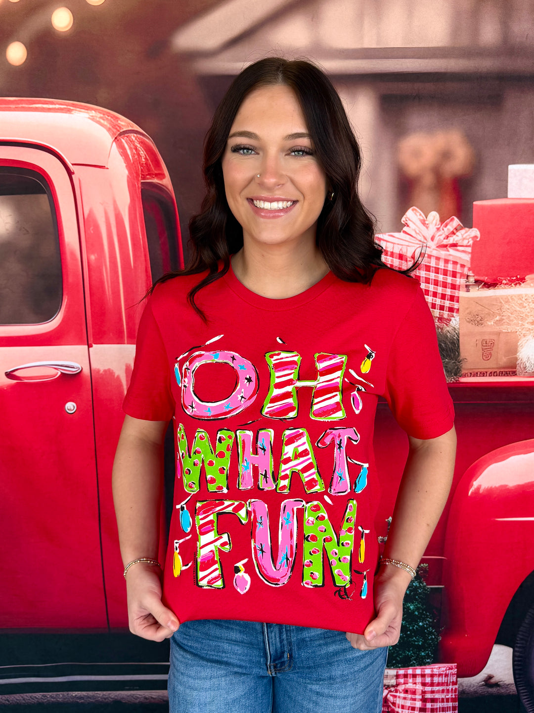 A woman wearing a bright red graphic t-shirt with the text 'OH WHAT FUN' in colorful, playful lettering, posed in front of a red vintage pickup truck with holiday decorations.