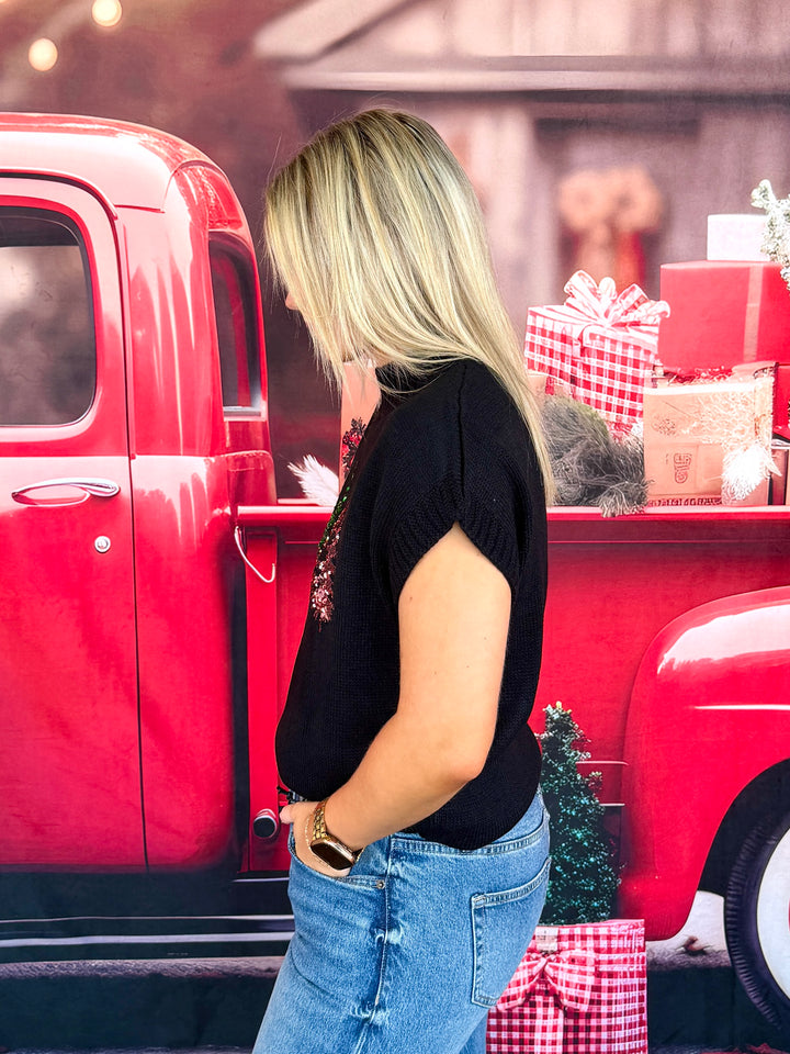 A woman with blonde hair is shown from the side, wearing a black short-sleeve knit top and blue jeans, standing next to a red vintage pickup truck decorated with holiday gifts.