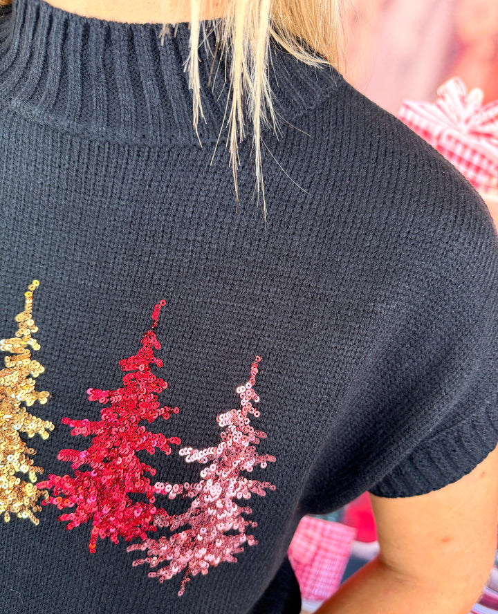 Close-up of a person wearing a black knit sweater with three sequined Christmas tree shapes in gold, red, and pink.