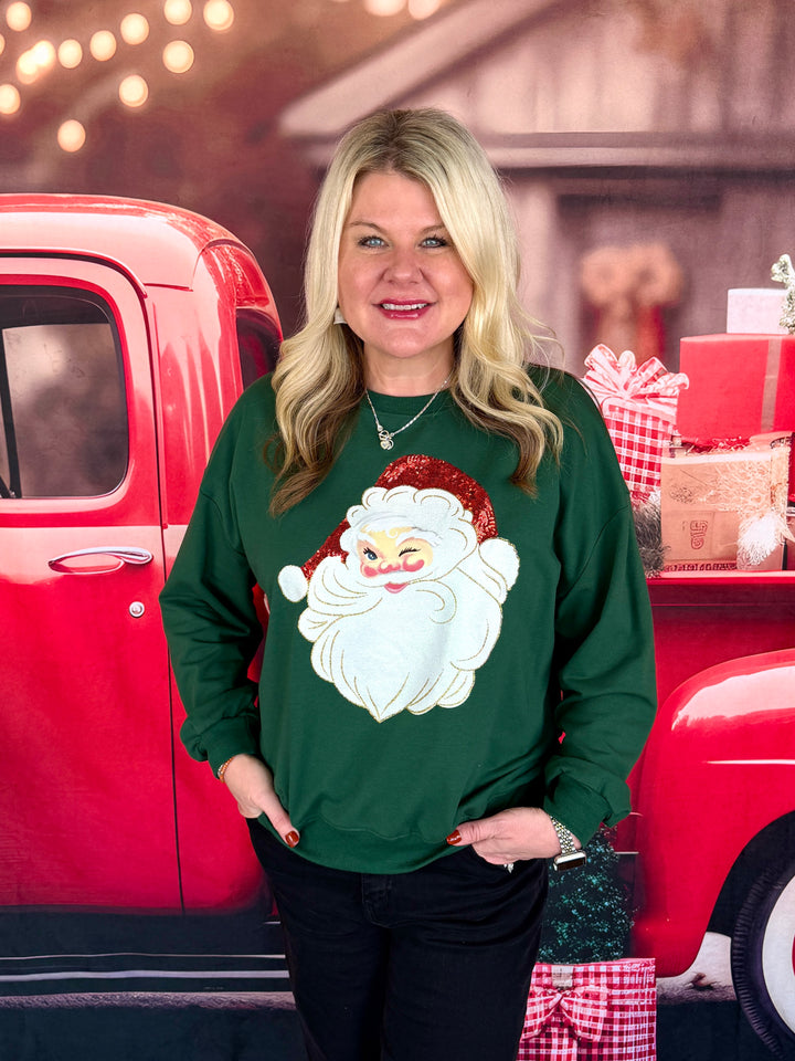 A woman wearing a dark green Christmas sweatshirt with a large white and red Santa Claus graphic on the front, standing in front of a red vintage truck with holiday decorations.