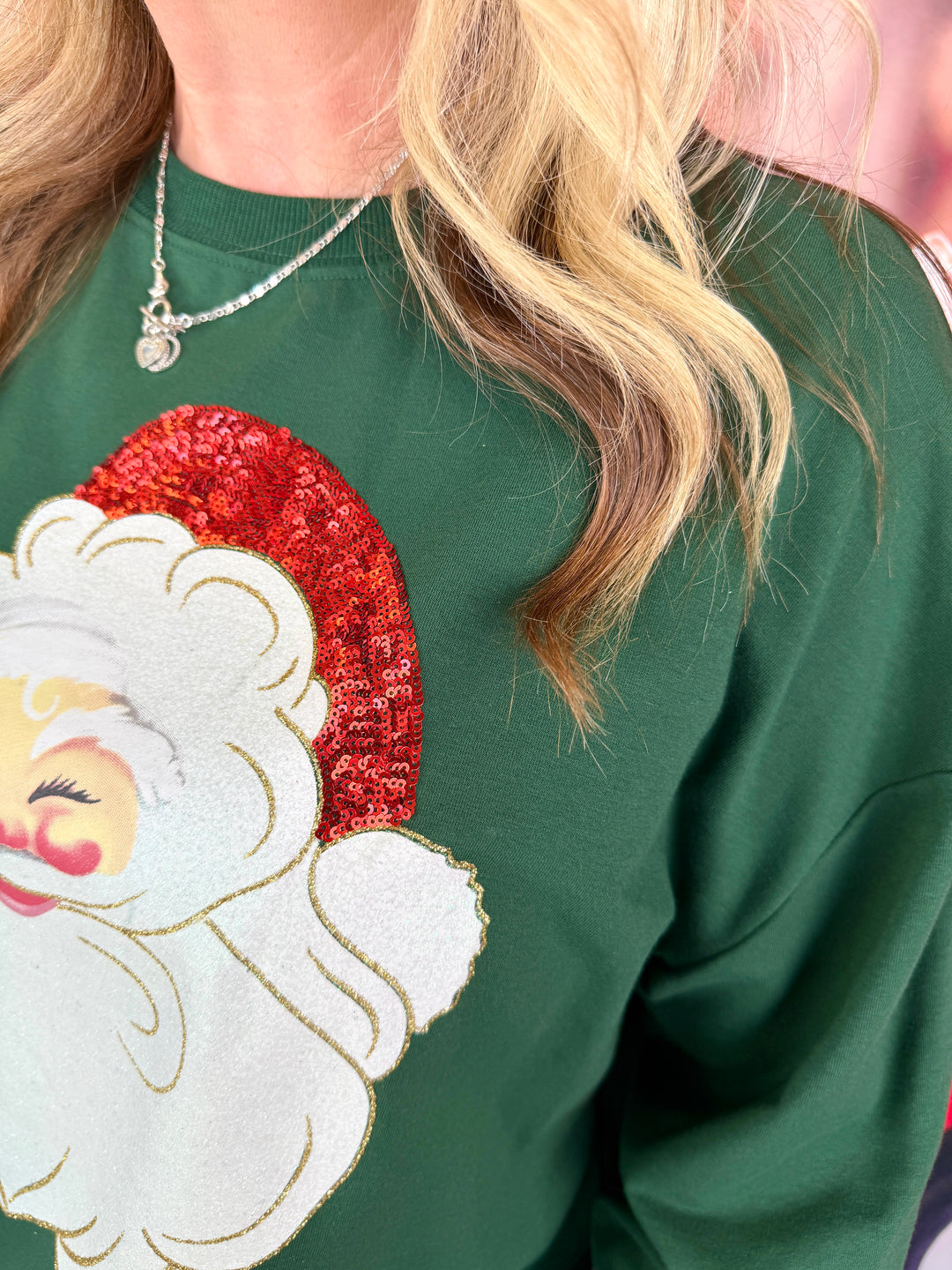 A close-up of a person wearing a dark green crewneck sweatshirt with a large sequined Santa Claus graphic on the front. The person has blonde hair and is wearing a silver necklace.