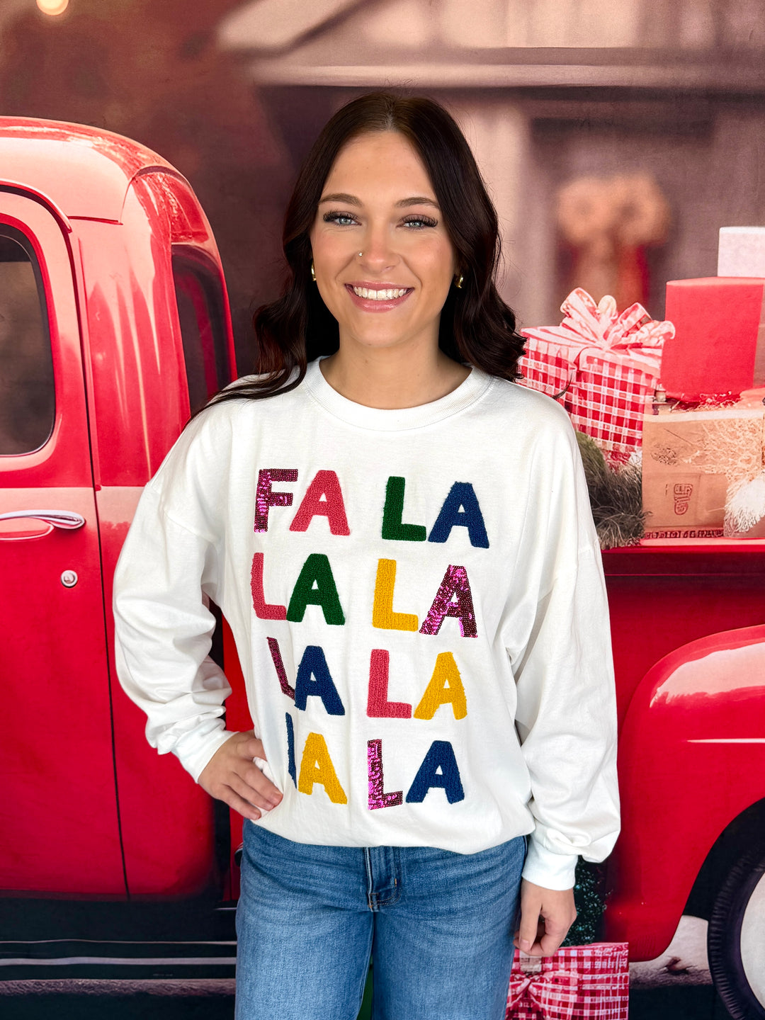 A woman smiling and wearing a white crewneck sweatshirt with 'FA LA LA LA LA LA LA LA LA' printed in colorful glittery letters, posed in front of a red vintage truck with holiday gifts.