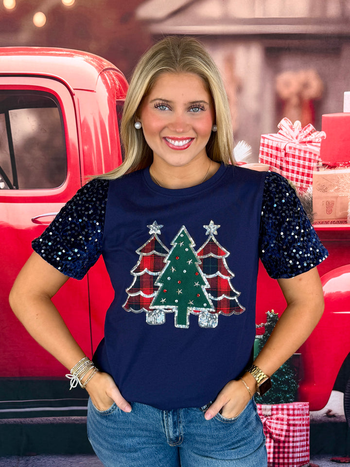 A woman wearing a navy blue Christmas-themed t-shirt with sequined puff sleeves and a tree graphic, posed in front of a red vintage truck backdrop.