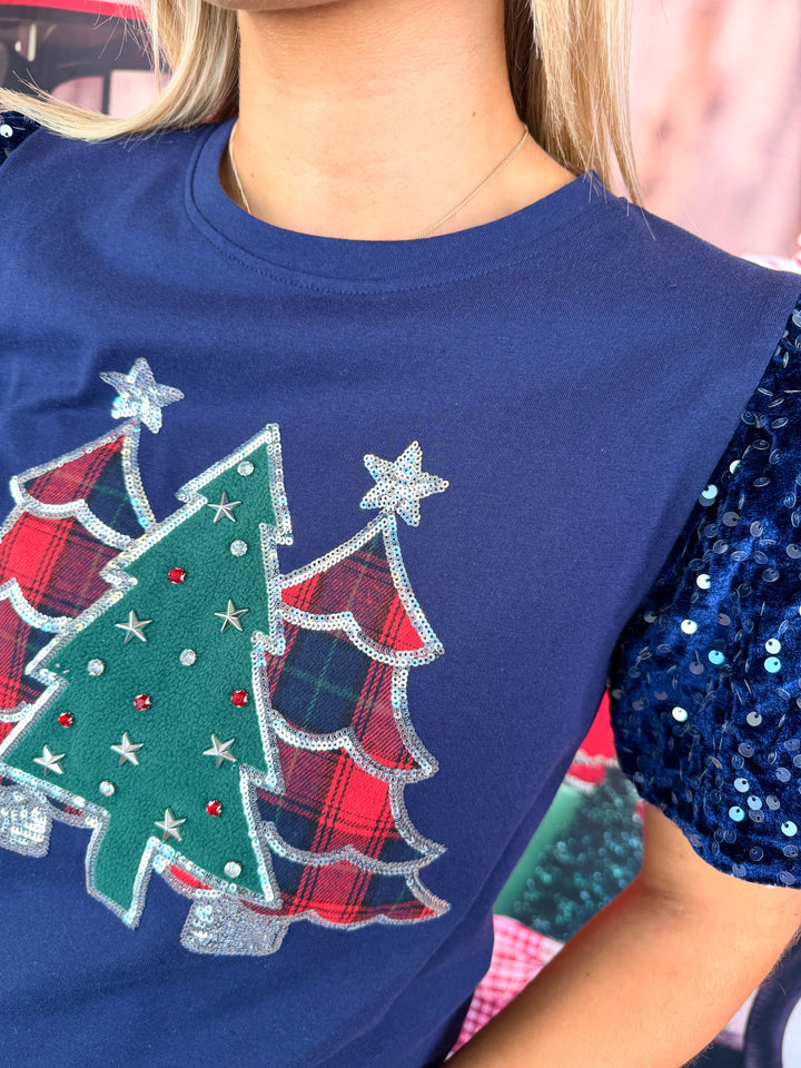 A close-up of a person wearing a navy blue holiday-themed sleeveless top with a sequined blue sleeve and a Christmas tree applique featuring green and red plaid trees with silver glitter trim.