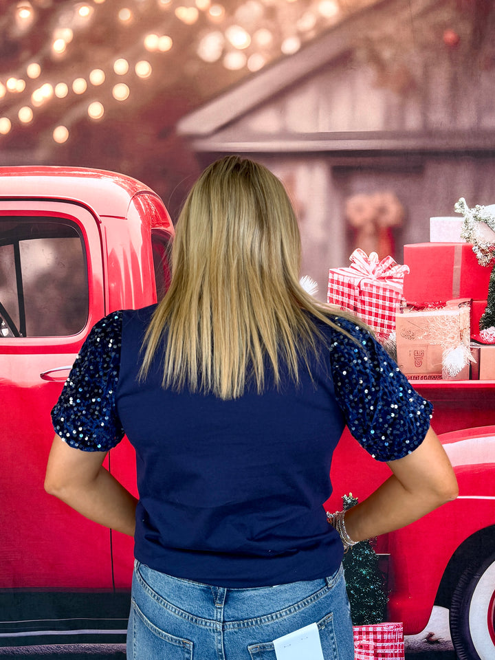 A woman viewed from behind wearing a navy blue top with sparkling silver sequin puff sleeves, standing in front of a red vintage pickup truck decorated with Christmas gifts.