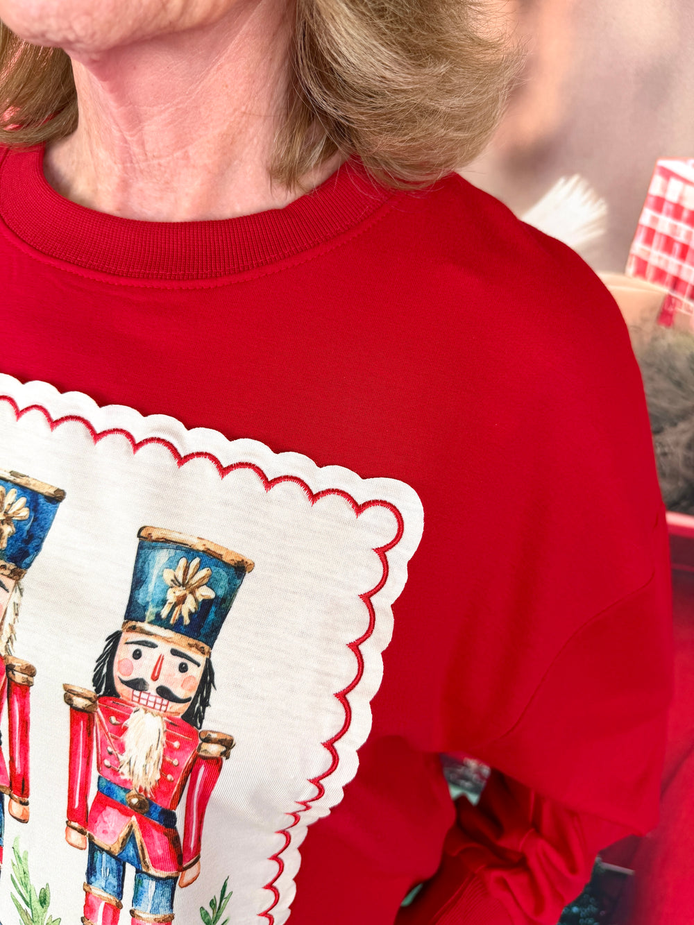 A close-up of a person wearing a bright red crewneck sweatshirt with a large white scalloped graphic panel featuring illustrated nutcracker figures.