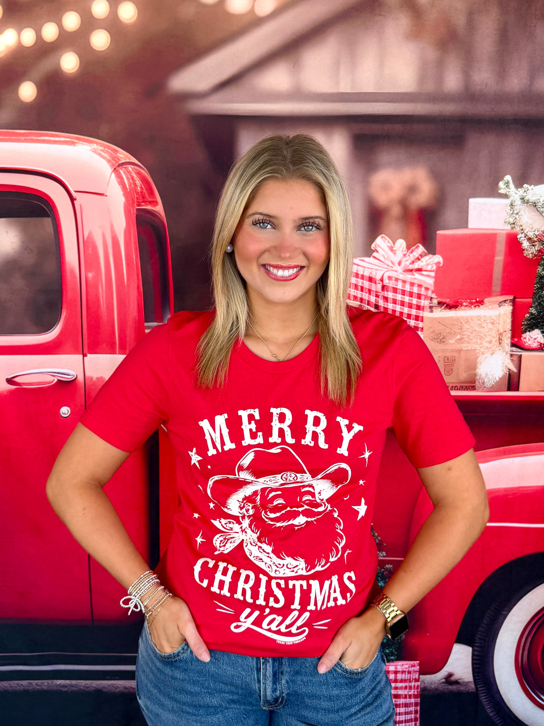 A smiling woman wearing a red graphic t-shirt with a white Santa Claus design and the text 'MERRY CHRISTMAS y'all', standing in front of a red vintage pickup truck with holiday decorations.