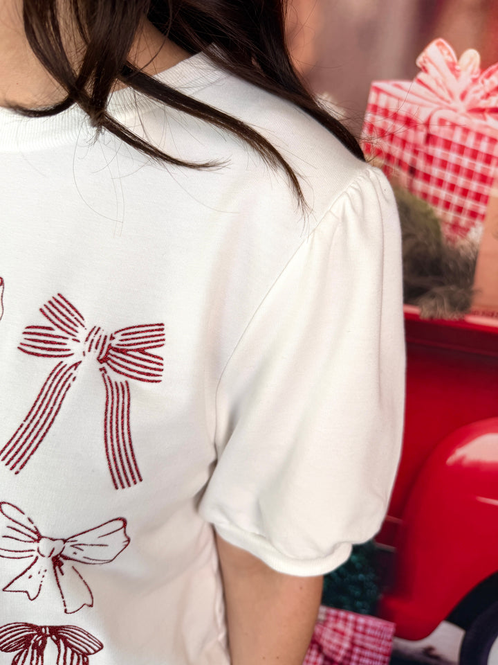 A close-up of a person wearing a white short-sleeve top with red ribbon and bow illustrations on the front, standing next to a red decorative object and wrapped gift boxes.