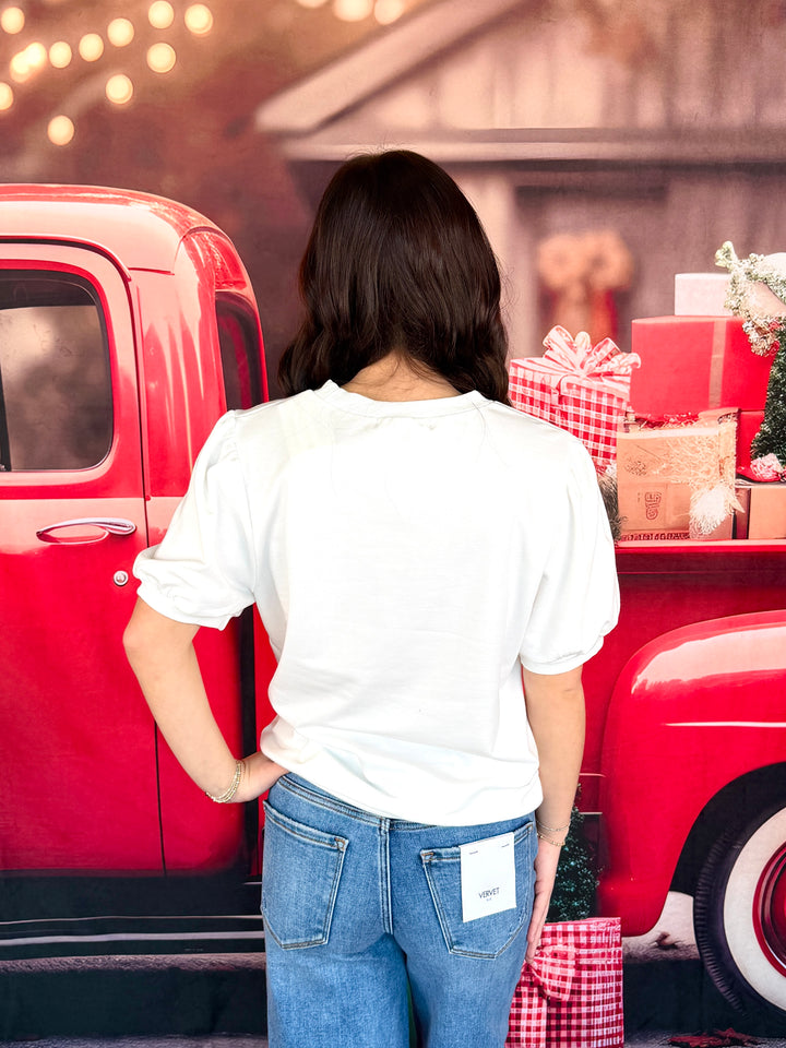 A woman viewed from behind wearing a white short-sleeve t-shirt and blue jeans, standing in front of a red vintage pickup truck decorated with holiday gifts.