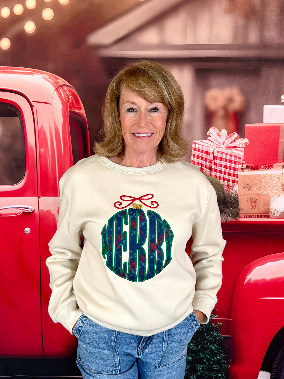 A woman wearing a cream-colored crewneck sweatshirt with a green and red 'MERRY' ornament graphic stands in front of a red pickup truck decorated for Christmas.
