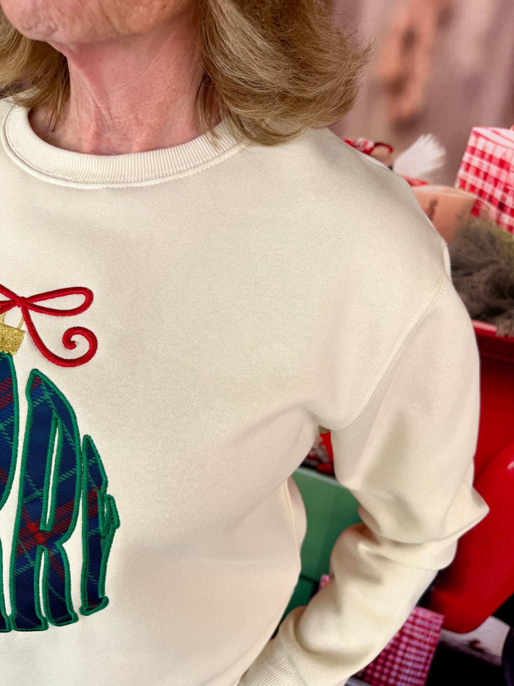 A close-up of a person wearing a cream-colored crewneck sweatshirt with a Christmas-themed graphic on the front, featuring green and red embroidered details.