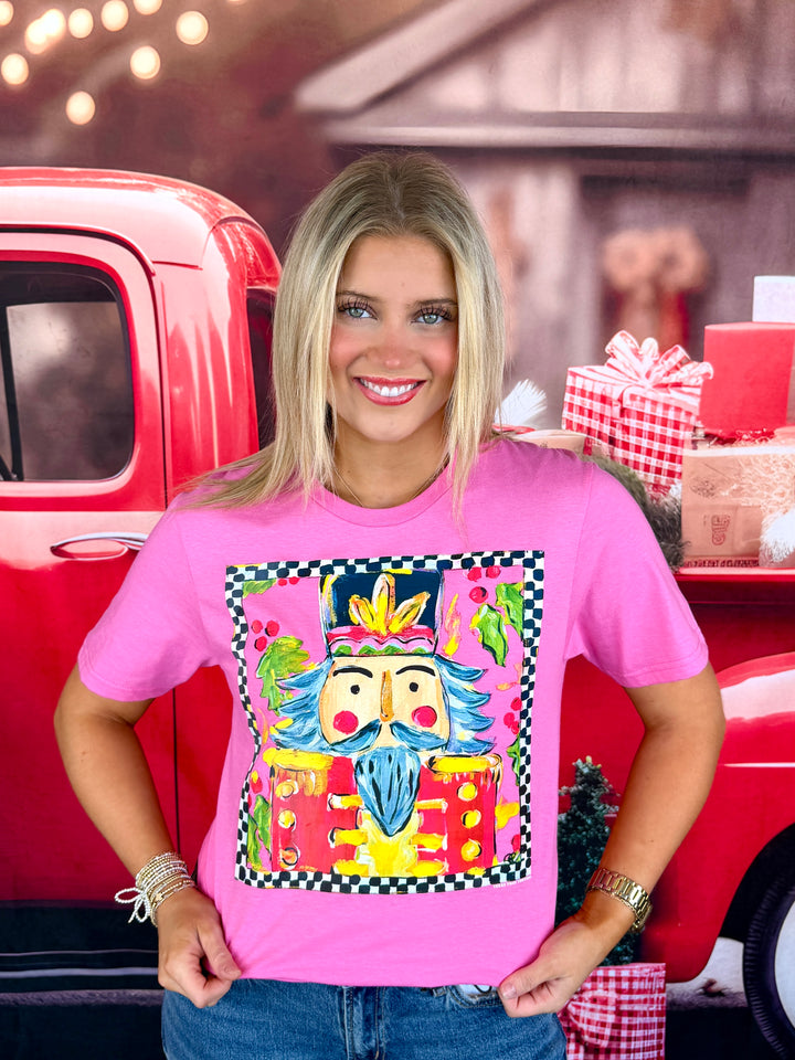 A smiling blonde woman wearing a bright pink graphic t-shirt with a colorful nutcracker illustration, posed in front of a red vintage truck and holiday decorations.