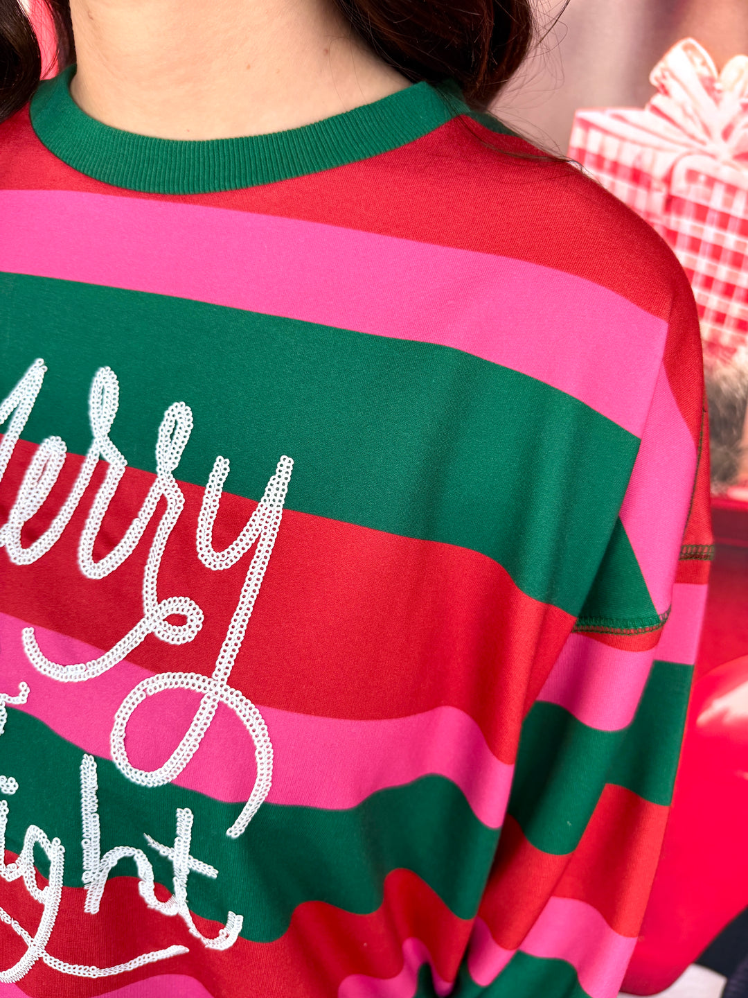 A close-up of a person wearing a festive holiday sweatshirt with horizontal stripes in red, pink, and green, featuring a white rhinestone-embellished 'Merry Night' script on the front.