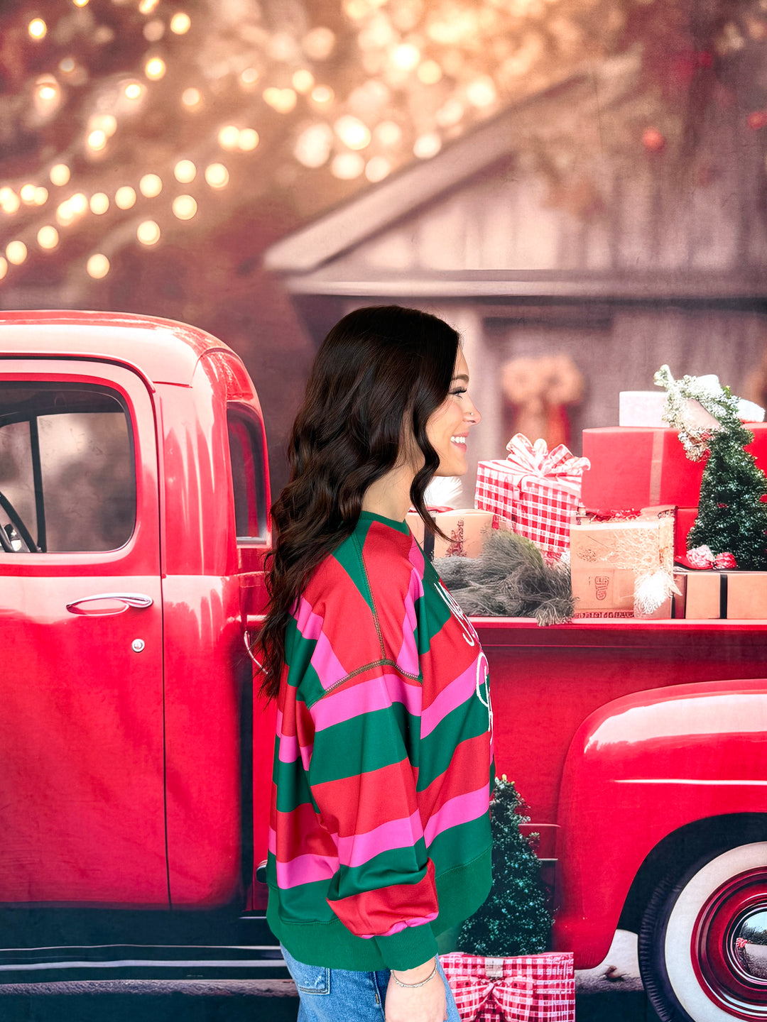 A woman wearing a red and green striped Christmas sweater stands next to a vintage red pickup truck loaded with holiday gifts and a small Christmas tree.