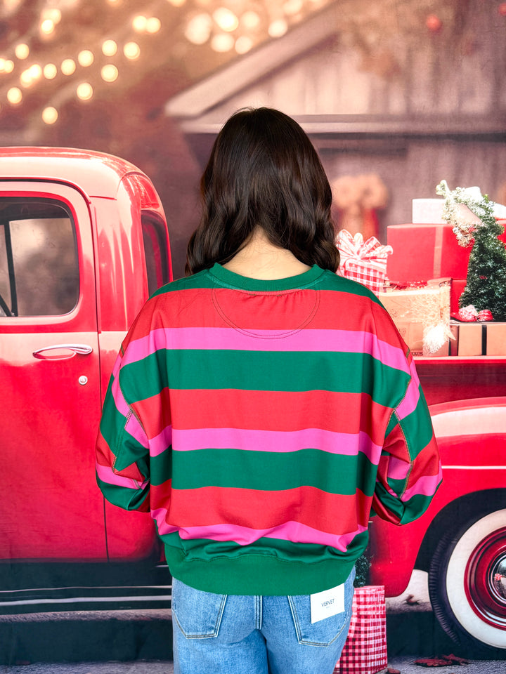 A woman viewed from behind wearing a festive holiday striped sweatshirt with horizontal bands of green, pink, and red, standing in front of a red vintage pickup truck decorated with Christmas gifts.