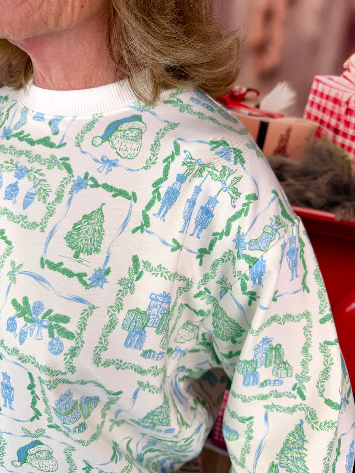 A close-up of a person wearing a white crewneck sweatshirt with a green and blue Christmas-themed toile print, set against a festive background with wrapped gifts.
