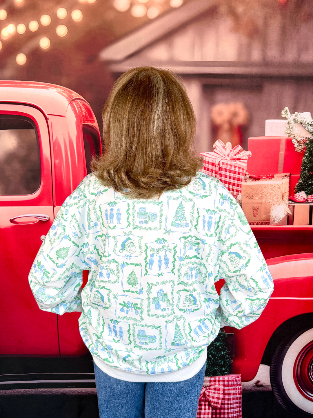 A woman viewed from behind wearing a white long-sleeve sweatshirt with a green and blue Christmas-themed pattern, standing in front of a red vintage pickup truck with wrapped gifts in the bed.