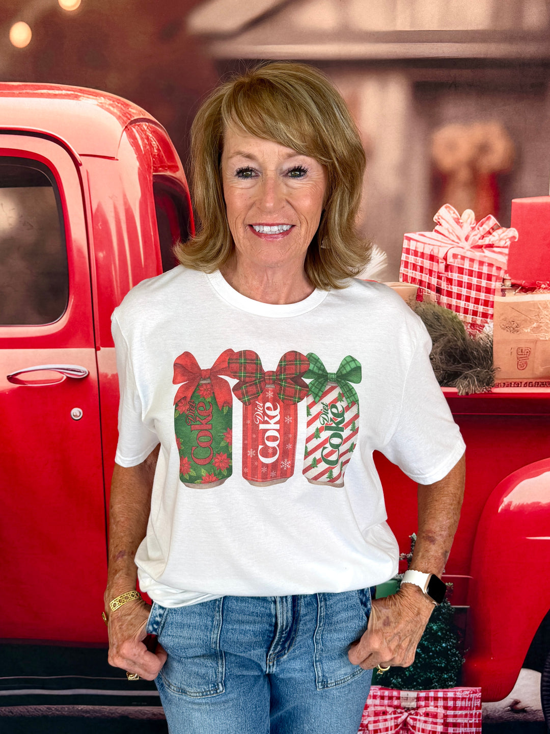 A woman wearing a white graphic t-shirt with a Christmas-themed Coca-Cola design, standing in front of a red vintage pickup truck with holiday gifts.