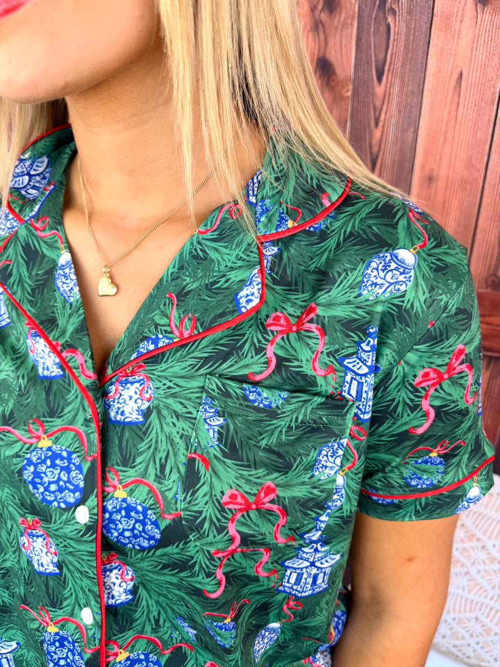 A close-up of a person wearing a short-sleeve pajama top with a Christmas tree print. The pajama shirt features a dark green background with pine branches, blue ornaments, and red ribbon accents, with red piping along the collar and placket.
