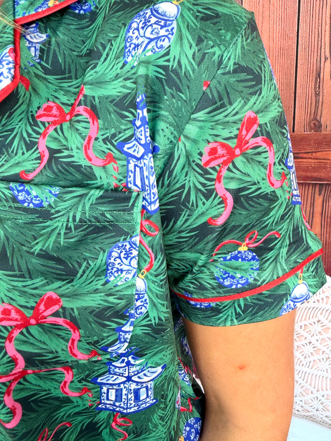 A close-up of a person wearing a short-sleeve button-up shirt with a Christmas tree print. The pattern features green pine needles, blue-and-white pagoda ornaments, and red ribbon bows, with red piping along the collar and sleeve cuffs.