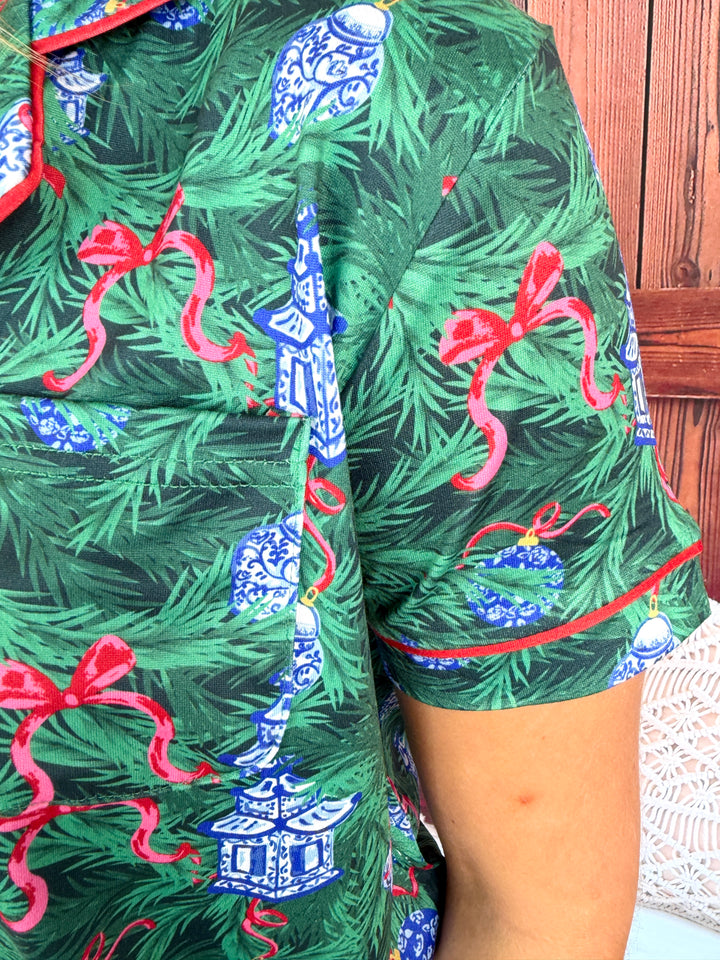 A close-up of a person wearing a short-sleeve button-up shirt with a Christmas tree print. The pattern features green pine needles, blue-and-white pagoda ornaments, and red ribbon bows, with red piping along the collar and sleeve cuffs.